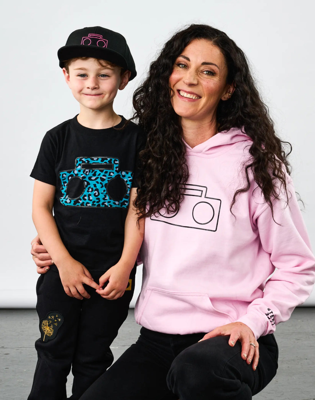Child wearing StereoType Kids Cheetah Boombox T-Shirt and Boombox Knit Sweatpants with pink Boombox Baseball Cap next to adult wearing pink Boombox Logo Hoodie