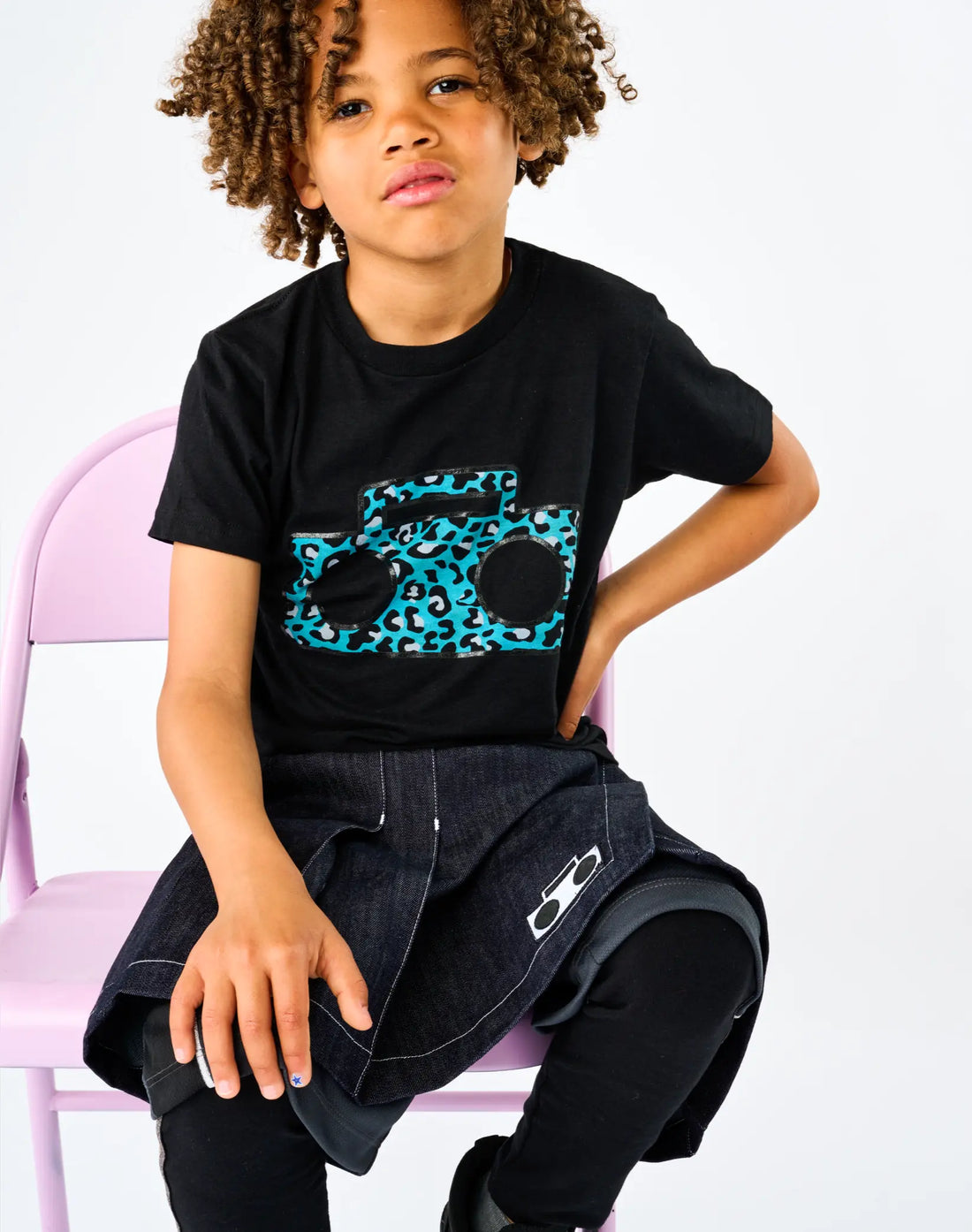 Child seated wearing StereoType Kids Cheetah Boombox unisex kids black graphic tee with Athletic Skort and Sparkle Leggings, gender-inclusive blended fashion made in the USA