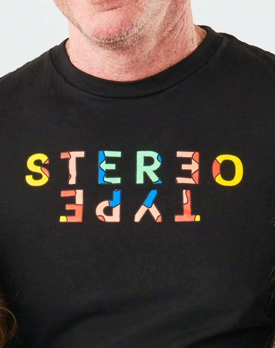 Close-up of StereoType Kids adult Kaleidoscope Logo Tee showing multicolor mirrored STEREOTYPE wordmark with reversed TYPE on black 100% cotton, screen printed in California