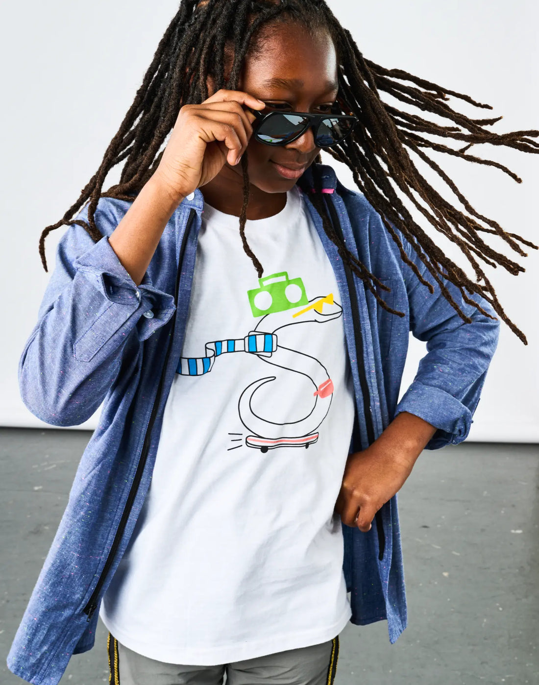Child with locs wearing StereoType Kids OG Stereo the Snake unisex kids white graphic tee layered under Chambray Button-Down Zip Shirt, sunglasses in hand