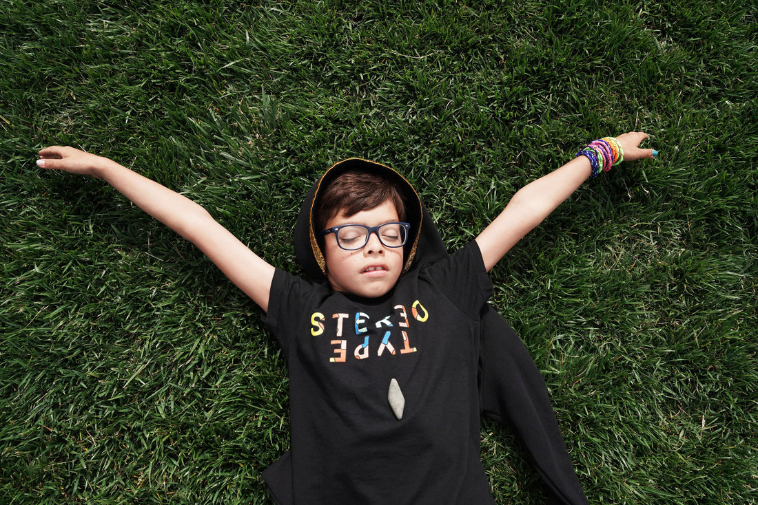 StereoType Kids co-founder Jacob lying in grass wearing the Kaleidoscope Logo hoodie tee, arms outstretched