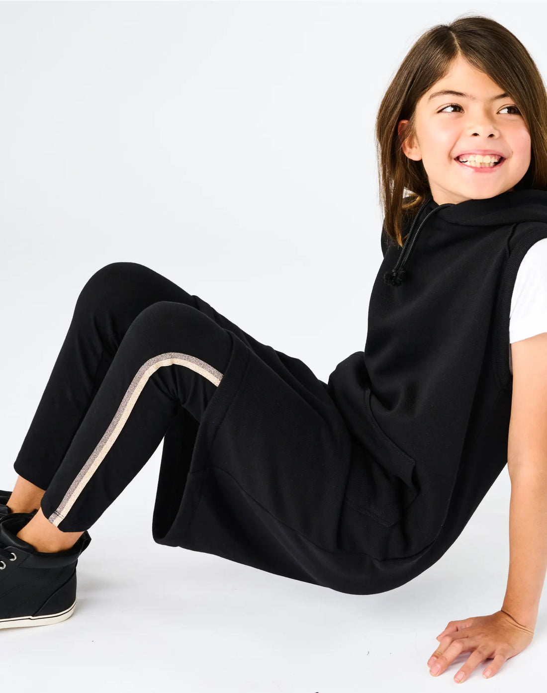 Chloe seated wearing StereoType Kids Sparkle Leggings in black with gold sparkle stripe, styled with black hoodie, unisex kids everyday leggings